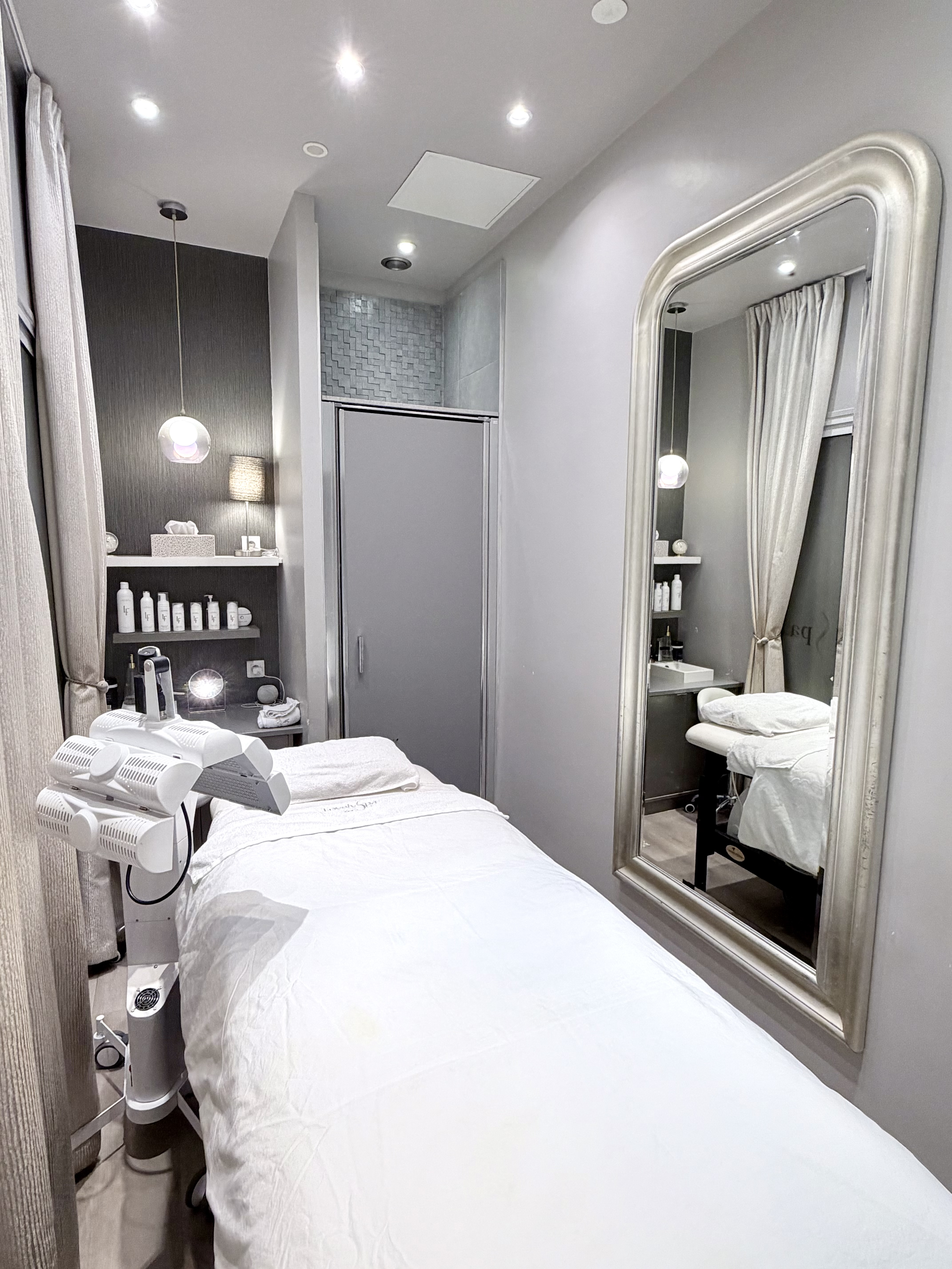 Intérieur d'une salle de soin avec un lit de massage, des produits de beauté sur une étagère et un grand miroir.